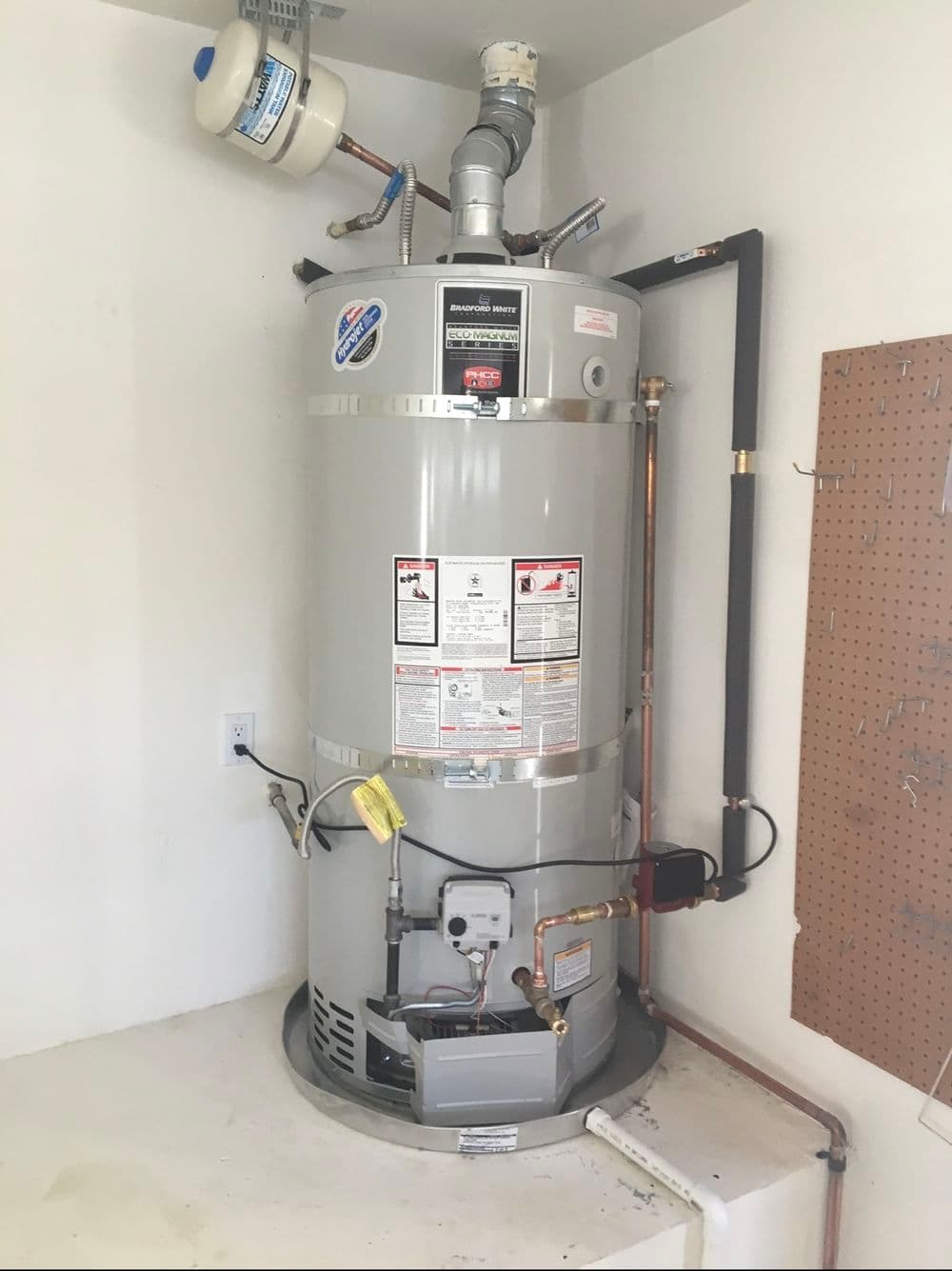Gray water heater installed in a home corner with visible plumbing and safety labels.
