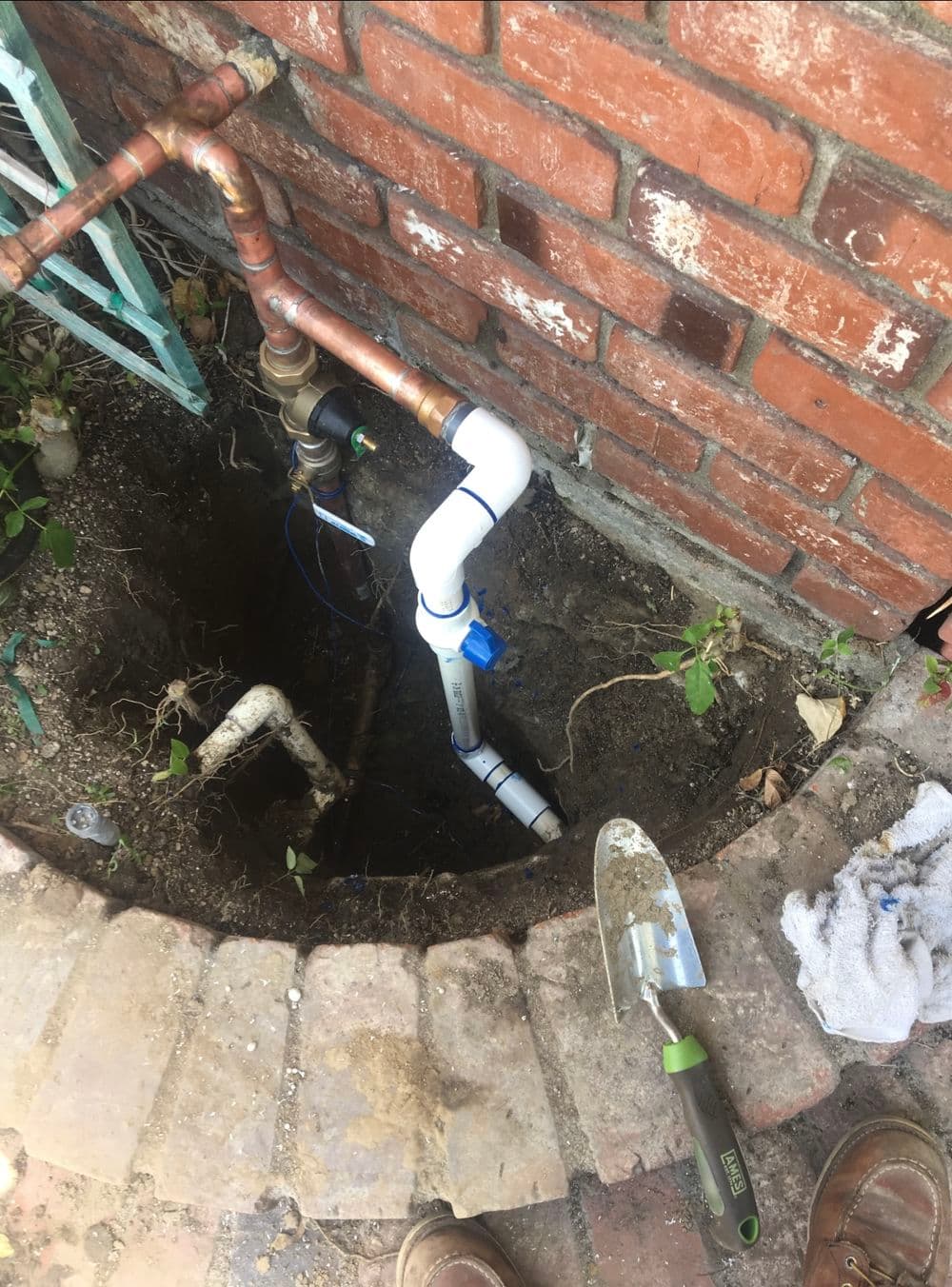 PVC pipe installation in a dug-out area near brick wall with gardening tools nearby.