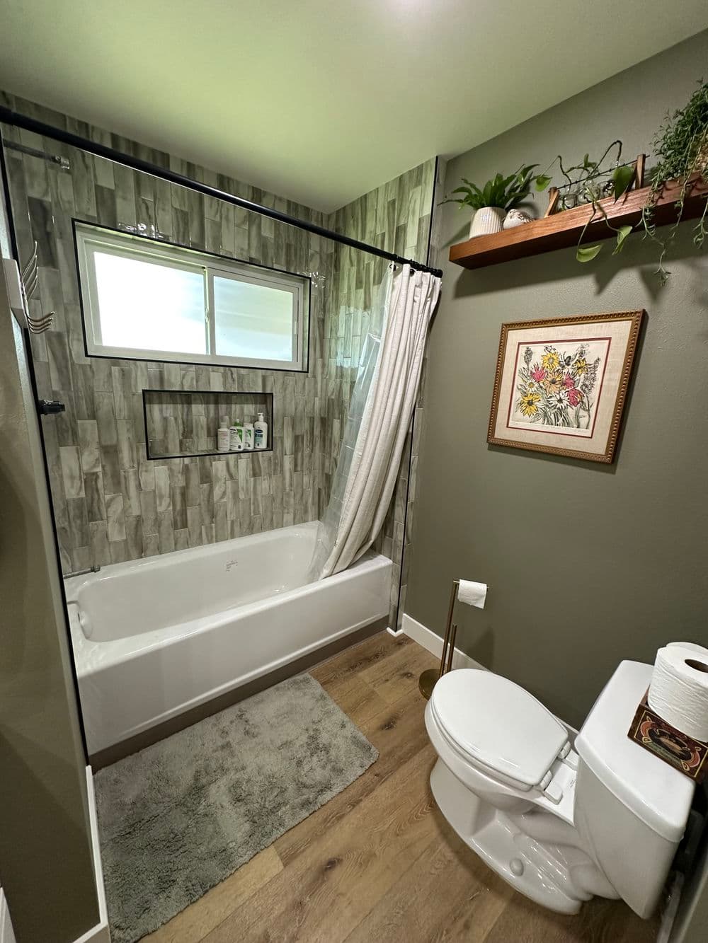 Modern bathroom with bathtub, glass shower, plants, and wooden shelf accents.
