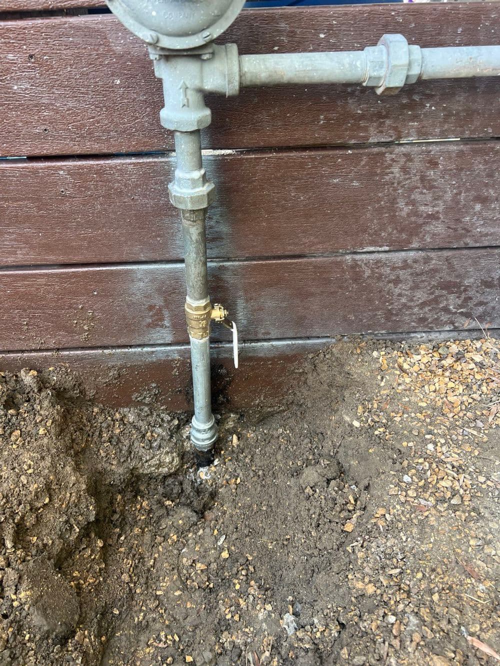 Exposed gas pipe connection with valve, installed near wooden fence and soil.