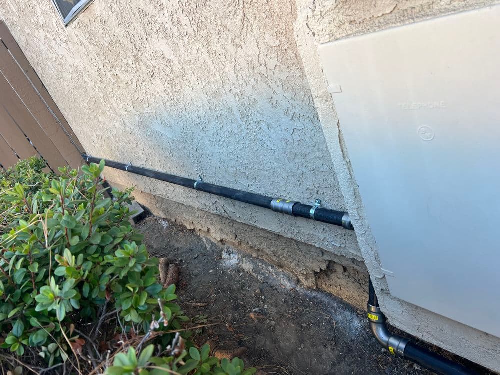Black plumbing pipe installed along a textured wall beside a utility box and greenery.