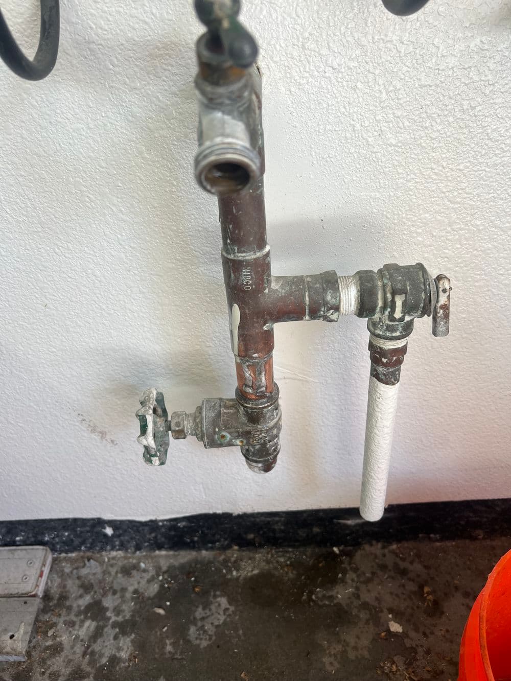 Rusty plumbing valve and pipes mounted on a textured wall, with a bucket nearby.