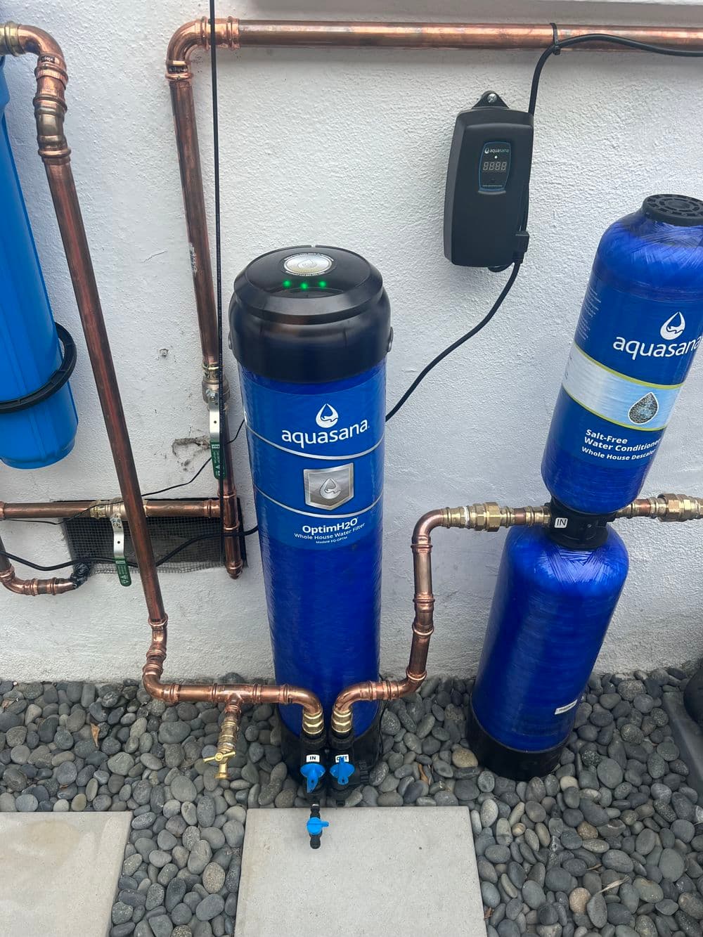 Aquasana water filtration system with blue casing and copper pipes, installed outdoors.