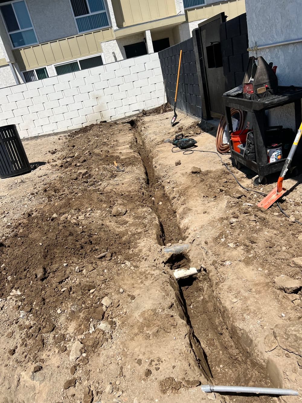 Excavated trench in a construction site with tools nearby and a building in the background.