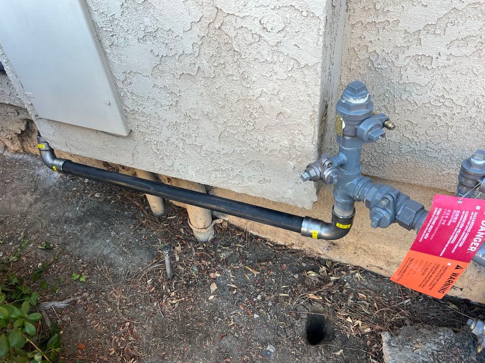 Gas meter installation with pipes and warning tag on exterior wall.