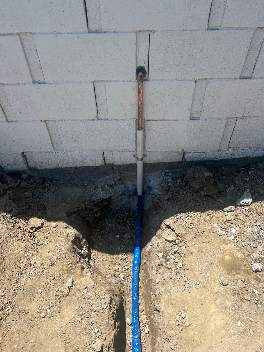 Copper and PVC pipe installation near a concrete block wall with exposed dirt.